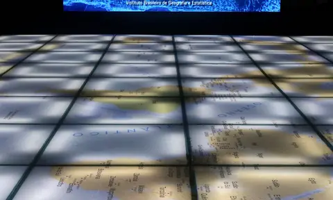Mapa-mundi iluminado no piso de uma sala do IBGE, com o nome do instituto num painel ao fundo